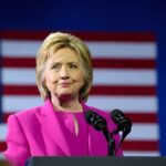 Person in pink suit speaking with American flag backdrop.