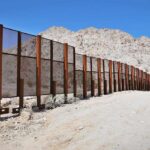 Border Wall Goes DARK – Literally! A long border wall stretches across a desert landscape with mountains in the background