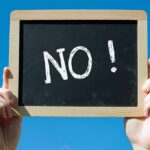 A hand holding a chalkboard with the word NO! written on it against a blue sky