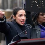 A woman passionately speaking at a rally with a sign in the background
