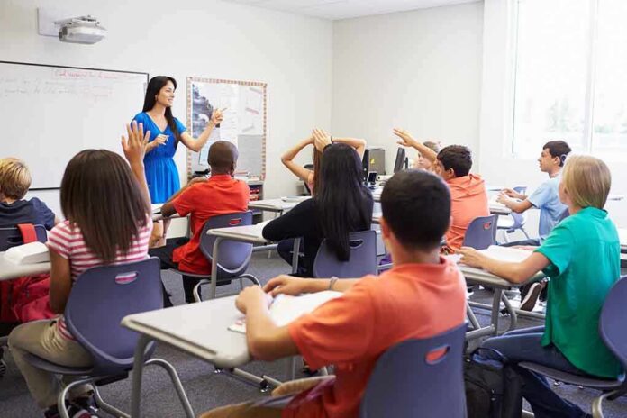 shutterstock_199144970.jpg Teacher in a blue dress instructing students in a classroom with hands raised