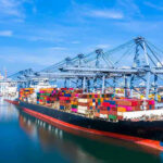 Iran Drones Hit Bahrain — War Crimes? Container ship docked at a busy industrial port.