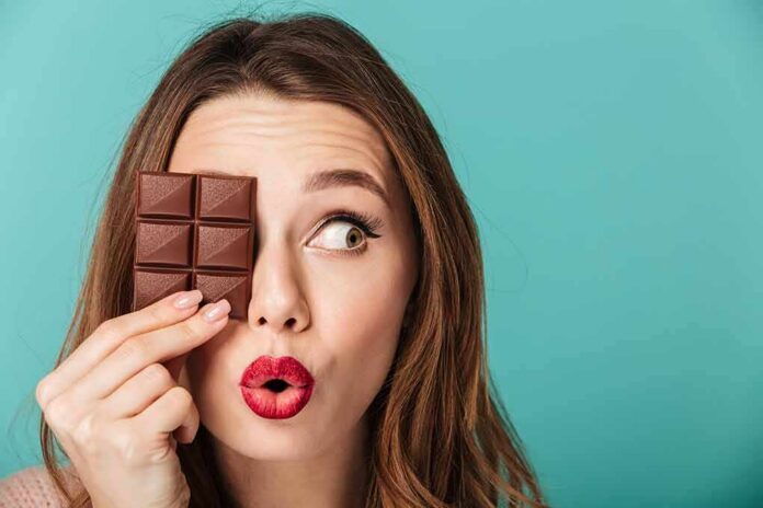 shutterstock_1007917828 Woman holding chocolate bar over one eye against teal background