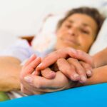Lowest Death Rate Ever — What’s Behind It? A caregiver holding the hands of an elderly patient in a hospital bed