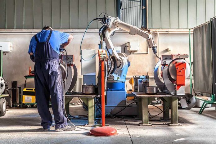 shutterstock_660540880 Factory worker operates near an industrial robot arm in a workshop