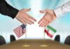 Two hands reaching to shake above US and Iran flags on a conference table