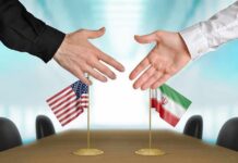 Trump’s Iran Ultimatum: Deal Or Annihilation? Two hands reaching to shake above US and Iran flags on a conference table