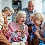 Later Retirement Wave Hits Hard Group of seniors smiling and looking at a smartphone together