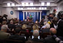 Trump’s Media Ban Turns Physical Press secretary at podium addressing reporters in briefing room