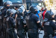 Classrooms Crumble, Streets Explode Riot police in helmets and visors stand in a line